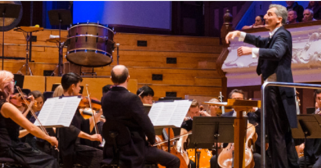 Um maestro regendo a Orquestra Filarmônica de Auckland em uma grande sala de concertos.