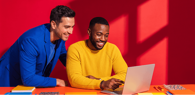 Two smiling men looking at a laptop on an orange desk, one in a blue blazer, the other in a yellow sweater, red background.