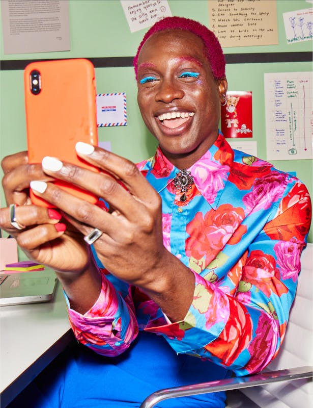 A person of colour with short pink hair, pink eyebrows, and bright blue eye shadow, holding a phone in front of them smiling.