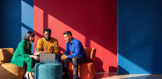 Three colleagues smile and collaborate around a laptop in a colorful modern lounge with red and blue walls.